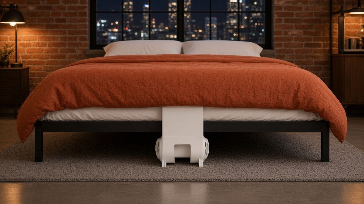 City loft bedroom at night with a rust orange duvet over a bFan under bed fan centered at the foot of a black metal bed